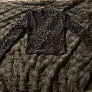 Glittrig svart långärmad topp - Säljer en svart glittrig långärmad topp i storlek XS. Toppen har en croppad passform och en snygg öppen rygg med knappstängning i nacken. Materialet är stretchigt och skimrar i glitter 