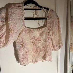 Blommig blus med puffärmar från H&M - Supersöt blommig blus från H&M i ljusbeige med rosa och gula blommor. Toppen har puffiga korta ärmar, smockad kropp och knytband vid axlarna. Perfekt för en somrig look med romantiska vibes.
