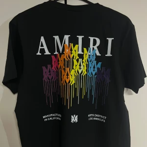 Svart AMIRI t-shirt med regnbågstryck - Svart t-shirt från AMIRI med stort regnbågsfärgat drip-tryck och logga på ryggen samt mindre tryck framtill. Rund hals och korta ärmar. Materialet är mjuk bomull och passformen är normal. Snygg statement-piece med färgstark design.