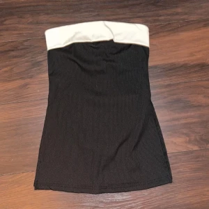 Svart tube top - Snygg svart tube top med vit kant upptill. Perfekt för en stilren look. Tröjan har en tight passform och är gjord i ett ribbat men skönt material. Den har även 2 slit i sidorna. Använt ett få tal gånger