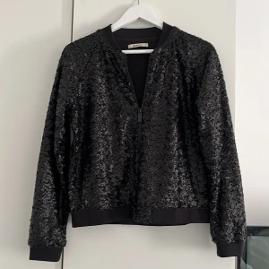 Svart paljett kofta Gina Tricot - Säljer en svart kofta från Gina Tricot, täckt med glittriga paljetter. Jackan har dragkedja framtill, ribbade muddar