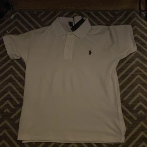 Vit Polo Ralph Lauren t-shirt slim fit - Vit t-shirt från Polo Ralph Lauren i custom slim fit-modell. Klassisk pikékrage med knappar och ikoniska broderade loggan på bröstet. Tillverkad i mjuk bomull som känns skön mot huden. Perfekt för en clean och stilren look.