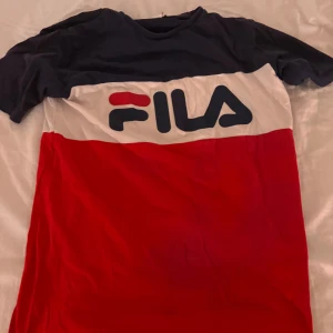 FILA t-shirt i rött, vitt och blått - Snygg FILA t-shirt med breda blockränder i marinblått, vitt och rött. Stor FILA-logga framtill. Klassisk rund hals och korta ärmar. Perfekt för dig som gillar sportig och street-inspirerad stil. Mjuk bomullskvalitet som är skön mot huden. Skickar bild på ifall det önskas. 