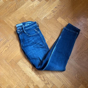 Blåa Replay anbass - Säljer ett par snygga Replay Anbass jeans i klassisk blå tvätt. Modellen är slim fit med raka ben och femficksdesign. Jeansen har tydliga sömmar, läderpatch bak och är tillverkade i mjukt denimtyg för en bekväm känsla.