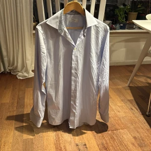 Ljusblå skjorta från Eton med mönstrad krage - Säljer en stilren ljusblå skjorta från Eton med snygga vita knappar. Insidan av krage och manschetter har ett diskret blåvitt mönster som ger extra edge. Skjortan är långärmad och i klassisk passform (Contemporary fit), perfekt för jobbet. Skjortan är i perfekt skick, använd en gång. En likadan skjorta , fast helt oanvänd finns här hemma och säljs i en annan annons på Plick