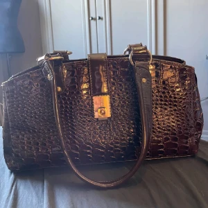 Mörkbrun handväska - Säljer denna fina väska då jag aldrig använder den, den är i väldigt bra skick då den typ aldrig är använd! Den är en blandning mellan mörkbrun och mörkröd.