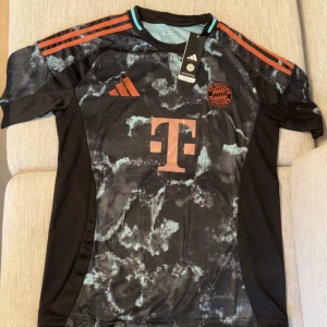 FC Bayern München svart matchtröja Adidas - Snygg FC Bayern München fotbollströja från Adidas med svart och turkos marmorerat mönster. Röd logga och ränder på axlarna, samt stor T-sponsor på bröstet. Kortärmad och tillverkad i lätt funktionsmaterial för bästa komfort på planen.