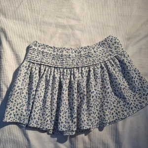 Blommig kortkjol från Gina Tricot - Supersöt kortkjol från Gina Tricot med blått blommönster på vit botten. Kjolen har smockad midja och är gjord i ett lätt, luftigt bomullstyg. Perfekt för en fräsch och somrig look! I strl 146-152 men passar om man har xxs eller xs ☺️