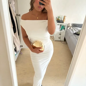 Vit offshoulder långklänning - Säljer en elegant vit långklänning med offshoulder-skärning och figurnära passform. Klänningen har snygga rynkade detaljer längs sidorna och är tillverkad i ett mjukt, stretchigt material som sitter perfekt. Perfekt för dig som vill sticka ut med en stilren look.