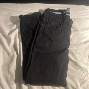 Svarta byxor från Jacob Cohën, stl 31 - Snygga svarta byxor från Jacob Cohën i storlek 31. Klassisk femficksmodell med silverfärgade detaljer och logga på bakfickan. Byxorna har normal passform och är tillverkade i ett mjukt bomullsmaterial med stilren look.