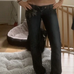 2000s Y2k low waist utsvängda Guess Premium jeans med broderi - Snygga mörkblå jeans från Guess Premium med broderade lövdetaljer på fickorna och kontrastsömmar. Klassisk femficksmodell med låg midja och utsvängda ben. Jeansen har slitningar och en unik patch bak i skinnimitation. Köpta second hand. Dom är i bra skick men lite slitna längst ner men inget man märker, kontakta mig så skickar jag fler bilder!