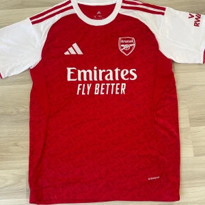 Arsenal röd kortärmad fotbollströja ( finns i L och M) - Snygg Arsenal fotbollströja från Adidas i rött och vitt med korta ärmar. Tröjan har klubbmärket på bröstet, Adidas-logga och Emirates Fly Better-tryck. Tillverkad i lätt och ventilerande material med subtilt mönster över hela framsidan.