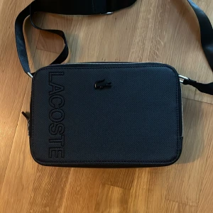 Lacoste axelväska  - Snygg svart axelväska från Lacoste med tydlig logga och krokodilemblem på framsidan. Pris och byte går att diskutera.