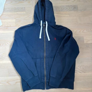 Mörkblå hoodie från Polo Ralph Lauren - Säljer en mörkblå hoodie från Polo Ralph Lauren med dragkedja och vit snörning i huvan. Klassisk röd logga broderad på bröstet och stora fickor framtill. Perfekt för dig som gillar en chill och stilren look.