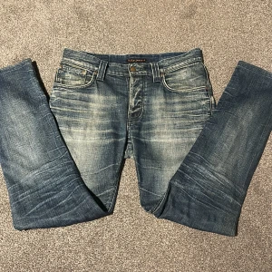 Nudie Jeans blå jeans 32/34 - Snygga blå jeans från Nudie Jeans i klassisk tvättad denim. Modellen har raka ben, fem fickor och coola slitningar framtill och baktill. Perfekt passform med normal midja och klassiska detaljer som läderpatch och kontrastsömmar.