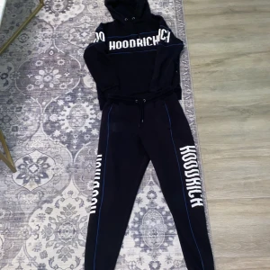 Svart hoodie från Hoodrich XS - Svart hoodie från Hoodrich i storlek XS med stor vit logga framtill och på ärmen. Tröjan har huva med snörning och blå detaljer längs sömmarna. Perfekt för dig som gillar streetwear och vill ha en snygg och bekväm look. Det är bara att skriva om ni vill köpa byxorna eller hoodien separat så tar vi priset sen