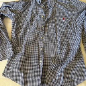 Rutig skjorta från Polo Ralph Lauren - Snygg blå och vit rutig skjorta från Polo Ralph Lauren i slim fit. Skjortan har klassisk krage, knäppning framtill och det ikoniska röda broderade logotypen på bröstet. Perfekt för dig som gillar stilrena och tidlösa plagg.
