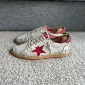 Golden Goose vita sneakers med röd stjärna - Golden Goose sneakers i vitt skinn med röd stjärna på sidan och röda detaljer vid häl och plös. Beige sula och slitna detaljer för en cool vintage-look. Klassisk rund tå, snörning och perforeringar framtill. Perfekta för dig som gillar streetstyle. Skorna är somsagt väldigt slitna därav det villiga priset. Skickar med dustbag.