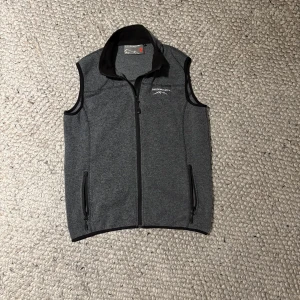 Grå fleeceväst från Swedemount - Mörkgrå fleeceväst från Swedemount med broderad logga på bröstet. Västen har hel dragkedja framtill, två fickor med dragkedja och hög krage. Perfekt att slänga över en t-shirt för en chill look.