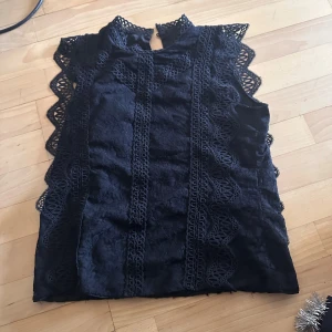 Svart spetsblus med volangdetaljer - Supersnygg svart blus med spets och volang längs ärmhålen och framstycket. Hög hals med knapp i nacken och vackra broderade detaljer. Perfekt för dig som vill ha en elegant och unik topp med coola detaljer.