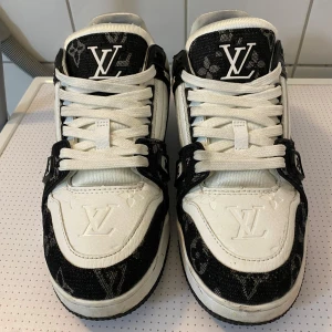 Louis vuitton skor - Snygga Louis Vuitton sneakers i svart och vitt med klassiskt monogrammönster. Skorna har rund tå, platt sula och är tillverkade i skinn och textil. Tjocka vita snören och tydliga LV-loggor framtill, på sidan och bak. Perfekta för dig som vill sticka ut med lyxig streetstyle.