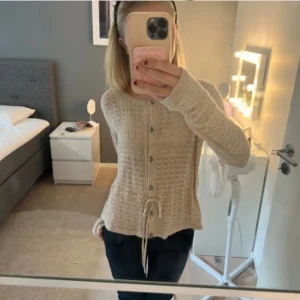 Beige stickad kofta med knappar - Superfin beige stickad kofta med snyggt mönster, knappar framtill och knytband i midjan. Långärmad och perfekt för lager-på-lager-stil. Passar dig som gillar en mysig och stilren look.