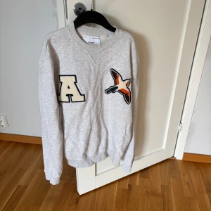 Grå sweatshirt Axel Arigato med patch Strl M - Grå sweatshirt från Axel Arigato med coola patches framtill – ett stort A och en broderad fågel i grönt, orange och brunt. Tröjan har rund hals, ribbade muddar och är långärmad. Perfekt för dig som gillar streetstyle och snygga detaljer. Storlek M men sitter mer som en S enligt mig!