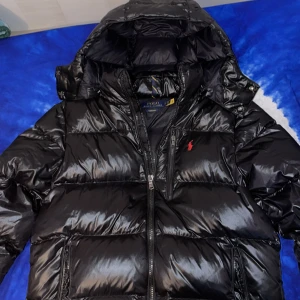 Svart pufferjacka Polo Ralph Lauren - Säljer en svart, glansig pufferjacka från Polo Ralph Lauren med huva och dragkedja. Jackan har klassisk quiltad design, två fickor med dragkedja och ikonisk röd logga på bröstet. Perfekt för kalla dagar när du vill hålla stilen.