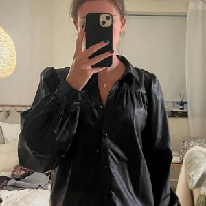 Svart skjorta Sofie Schnoor PU - Snygg svart skjorta från Sofie Schnoor med glansig yta i PU-material. Modellen har puffiga långärmar, klassisk krage och knappar framtill. Skjortan har en loose passform och coola detaljer vid axlarna som ger extra attityd.