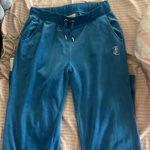 Blå velourbyxor från Juicy Couture - Blåa mjukisbyxor i velour från Juicy Couture med snörning i midjan och fickor. Fram finns en liten JC-logga i strass och på baksidan står det 'Juicy' i stora strasstenar. Perfekta för en chill och trendig look.