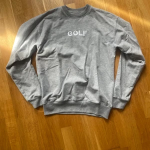 Grå crewneck från Golf Wang - Säljer en stilren grå crewneck från Golf Wang med broderad vit GOLF-text på bröstet. Tröjan är i mjuk bomulls- och polyestermix, har rund hals och långa ärmar. Perfekt för dig som gillar enkel streetwear med clean vibe.