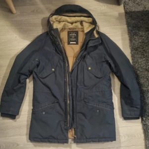 Mörkblå parkajacka Jack & Jones - Parkajacka från Jack & Jones i mörkblått med varm beige foder och mysig teddykrage. Jackan har flera fickor med knappar, huva och dragkedja framtill. Perfekt för kalla dagar när du vill hålla stilen och värmen.