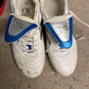 Vita inomhus fotbollsskor strl 43 - Säljer ett par vita inomhus fotbollsskor från Diadora med blå och gula detaljer. Skorna har snörning, gummisula i brunt och är tillverkade i syntetmaterial. Perfekta för futsal eller inomhusträning. Klassisk design med sportig känsla.