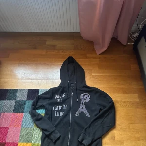 Svart hoodie med strass och Paris-tryck - Svart hoodie från BLINGLUCID med dragkedja, huva och snörning. Framsidan har strassdetaljer med texten 'paris au clair de lune' och motiv av Eiffeltornet och månen. Perfekt för dig som gillar statement-plagg med bling.