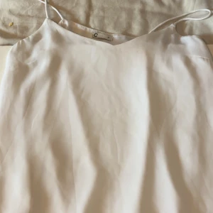 Vit kort klänning med smala axelband - Säljer en stilren vit klänning från C i storlek 34. Klänningen har smala axelband och en enkel, rak passform som ger en clean look. Materialet är mjukt och har en lätt glans, vilket gör den extra fräsch. Perfekt för dig som gillar minimalistisk stil.