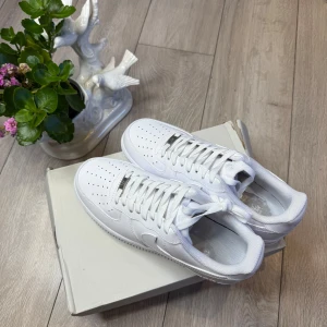 Nike Air Force 1 helvita sneakers - Klassiska Nike Air Force 1 i helvitt skinn med perforerad tå, rund tå och platt sula. Snygg och tidlös modell med snörning och diskret Nike-logga på sidan och hälen. Perfekta för dig som vill ha en clean och stilren look. ✅ Storlek 43 ✅ Helt nya, oanvända ✅ Skickas inom 1–3 dagar 