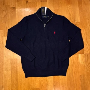 Mörkblå stickad half zip från Polo Ralph Lauren - Snygg mörkblå stickad tröja från Polo Ralph Lauren med half zip och klassisk röd broderad logga på bröstet. Tröjan har lång ärm, ribbade muddar och hög krage. Perfekt för dig som gillar stilren och tidlös design.