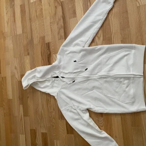 Vit hoodie från Polo Ralph Lauren - Snygg vit hoodie från Polo Ralph Lauren med dragkedja och klassisk broderad logga på bröstet. Tröjan har huva med snörning, två fickor fram och är tillverkad i mjuk bomull. Perfekt för en clean och stilren look.
