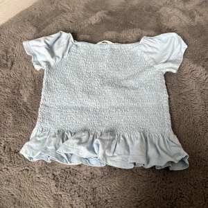 Ljusblå volangblus från H&M - Supersöt ljusblå blus från H&M med puffiga korta ärmar och smockad kropp. Nedtill finns en bred volangkant som ger en gullig och trendig look. Perfekt för dig som gillar romantiska detaljer och mjuka pasteller.