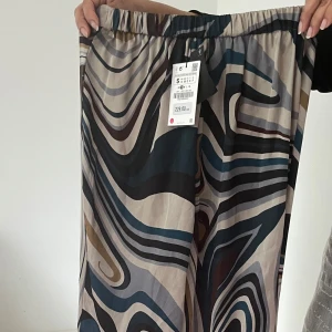Mönstrade vida byxor från Zara - Säljer ett par sjukt snygga vida byxor från Zara med abstrakt mönster i beige, grönt, svart och brunt. Byxorna har resår i midjan och är gjorda i ett tunt, flowy material som känns riktigt skönt