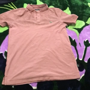 Rosa t-shirt från Lacoste med knappar - Säljer en rosa t-shirt från Lacoste med korta ärmar och klassisk ståkrage med knappar framtill. T-shirten har diskret strukturmönster upptill och den ikoniska krokodilloggan på bröstet. Tillverkad i mjuk bomull för en skön känsla.