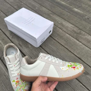 Maison Margiela sneakers med färgstänk - Säljer ett par vita Maison Margiela sneakers med färgstänk i grönt, rött, gult och blått. Skorna har en klassisk siluett med beige detaljer, snörning och gummisula. Tillverkade i skinn och mocka för en lyxig känsla. Box medföljer.