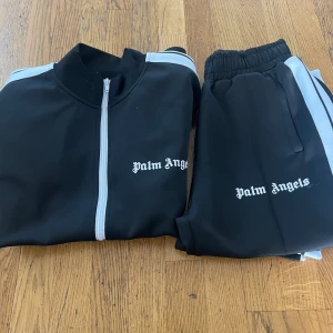 Palm Angels svart tracksuit med vita ränder - Säljer en svart tracksuit från Palm Angels med vita ränder längs ärmar och ben. Setet har dragkedja på jackan och elastisk midja på byxorna. Palm Angels-logga broderad på både jacka och byxor. Tillverkad i mjuk polyester för en sportig vibe.