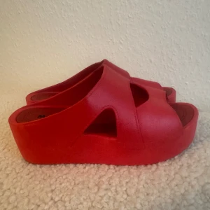 Röda platåsandaler med öppen tå - Coola röda sandaler med platåsula och öppen tå. Skorna har breda remmar med utskurna detaljer och en chunky plattform som ger extra höjd. Perfekta för dig som vill sticka ut med färg och stil. Tillverkade i syntetmaterial. Använd en gång. Innelängd 22,5cm, höjd 5cm.