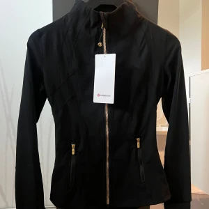 Svart define jacka från Lululemon - Svart, figursydd vindjacka från Lululemon med snygga guldiga dragkedjor och hög krage. Jackan har långa ärmar och två fickor framtill med dragkedja. Perfekt för dig som gillar en clean och sportig look. Passar storlekar xs-m. Kan också byta mot en annan färg 