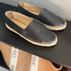 Svarta espadrilles från Chanel - Chanel espadrilles i svart skinn med klassisk CC-logga på ovansidan. Snygg flätad sula i naturfärgad jute och kontrastsömmar runt om. Perfekt för dig som vill ha en lyxig och stilren sko till sommaren. Original kvitto, kartong, dustbag, alla kort och snöre medföljer. 