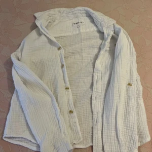 Vit bomullsskjorta från DMD för barn  - Luftig och stilren vit skjorta från DMD i mjuk, krinklad bomull. Skjortan har klassisk krage, knappar framtill och långa ärmar med knapp vid manschetten. Perfekt för en avslappnad och trendig look i storlek 9 år/ 134 cm. 