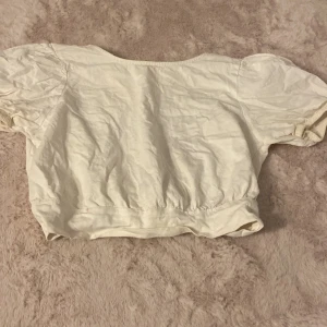 Vit croppad blus med knyt i ryggen - Supersöt vit croppad blus med korta puffärmar och snygg knytning i ryggen. Toppen har en öppen ryggdetalj och resår i midjan för en skön passform. Perfekt för dig som gillar romantisk och trendig stil.