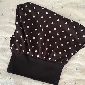 Svart prickig blus med bred mudd - Snygg svart blus med vita prickar och bred, enfärgad mudd nedtill. Toppen har en lös passform upptill och är ärmlös, perfekt för en trendig look. Materialet känns mjukt och följsamt, vilket gör den bekväm att bära hela dagen.