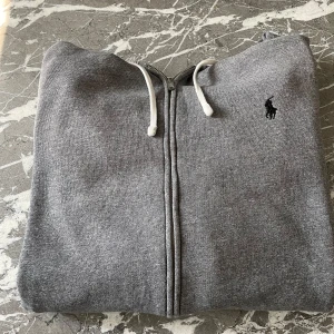 Ralph Lauren Zip Hoodie  - Mycket snygg och fräsch Zip Hoodie från Ralph Lauren k färgen grå. Hoodien är använd men fortfarande i ett bra skick. Däremot finns det ett pyttelitet håll på sidan av axeln men är analogt inte något som märks då man inte kollar jättenoga. Om du har några frågor kring produkten eller priset så är det bara att höra av dig.😄 Mvh Stockholm Wardrobe 